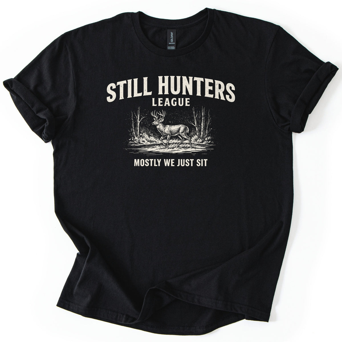 Still Hunters League Comfort Colors T-shirt in Black, laid flat. Cream text reads Still Hunters League above a sketch of a buck walking by water between bare trees, with Mostly We Just Sit below. The shirt has rolled sleeves on a white background.