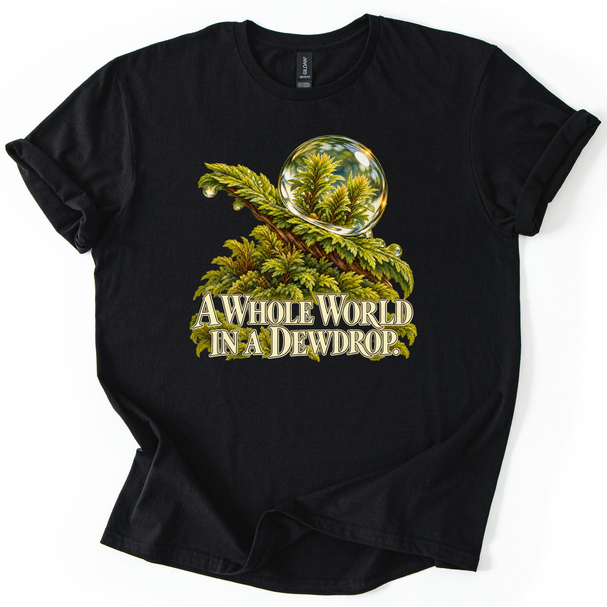 A Whole World in a Dewdrop Comfort Colors T-shirt in Black, laid flat. A large clear water droplet sits on a mossy green frond dotted with smaller drops, reflecting tiny plants, over cream text reading A Whole World in a Dewdrop. Sleeves rolled on a white background.