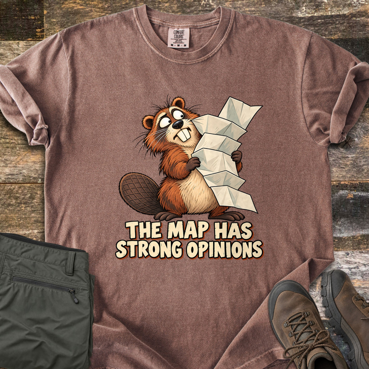 The Map Has Strong Opinions Comfort Colors T-shirt in Espresso, laid flat. A cartoon beaver with wide, crossed eyes clutches a tall, unfolding paper map above the words The Map Has Strong Opinions. Sleeves are rolled, beside green hiking pants and brown hiking boots on weathered wood.