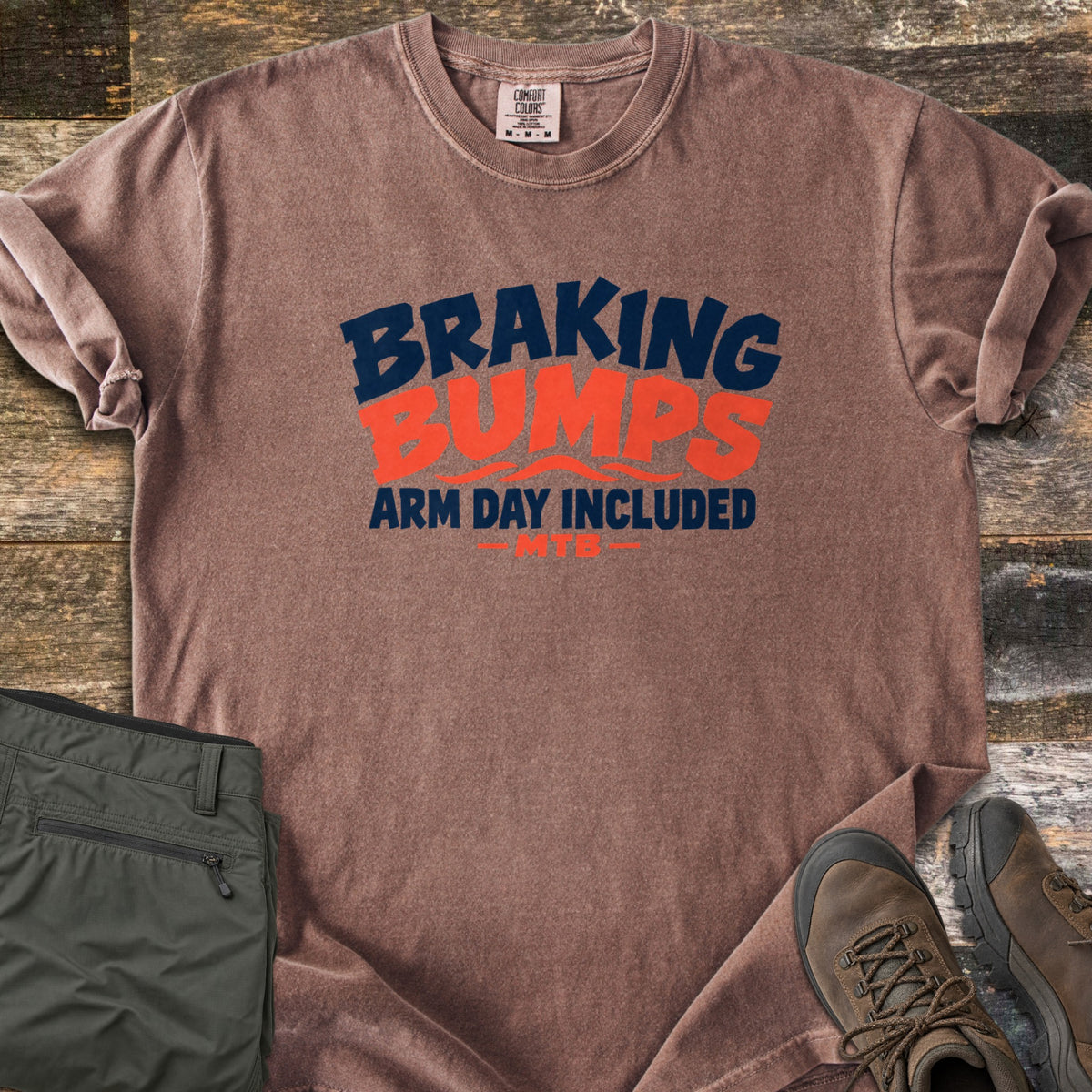 Braking Bumps Arm Day Included Comfort Colors T-shirt in Espresso, laid flat. Bold navy and orange text reads Braking Bumps, Arm Day Included, MTB, with wavy orange lines under Bumps. It rests on a wood floor beside olive cargo pants and brown hiking boots.