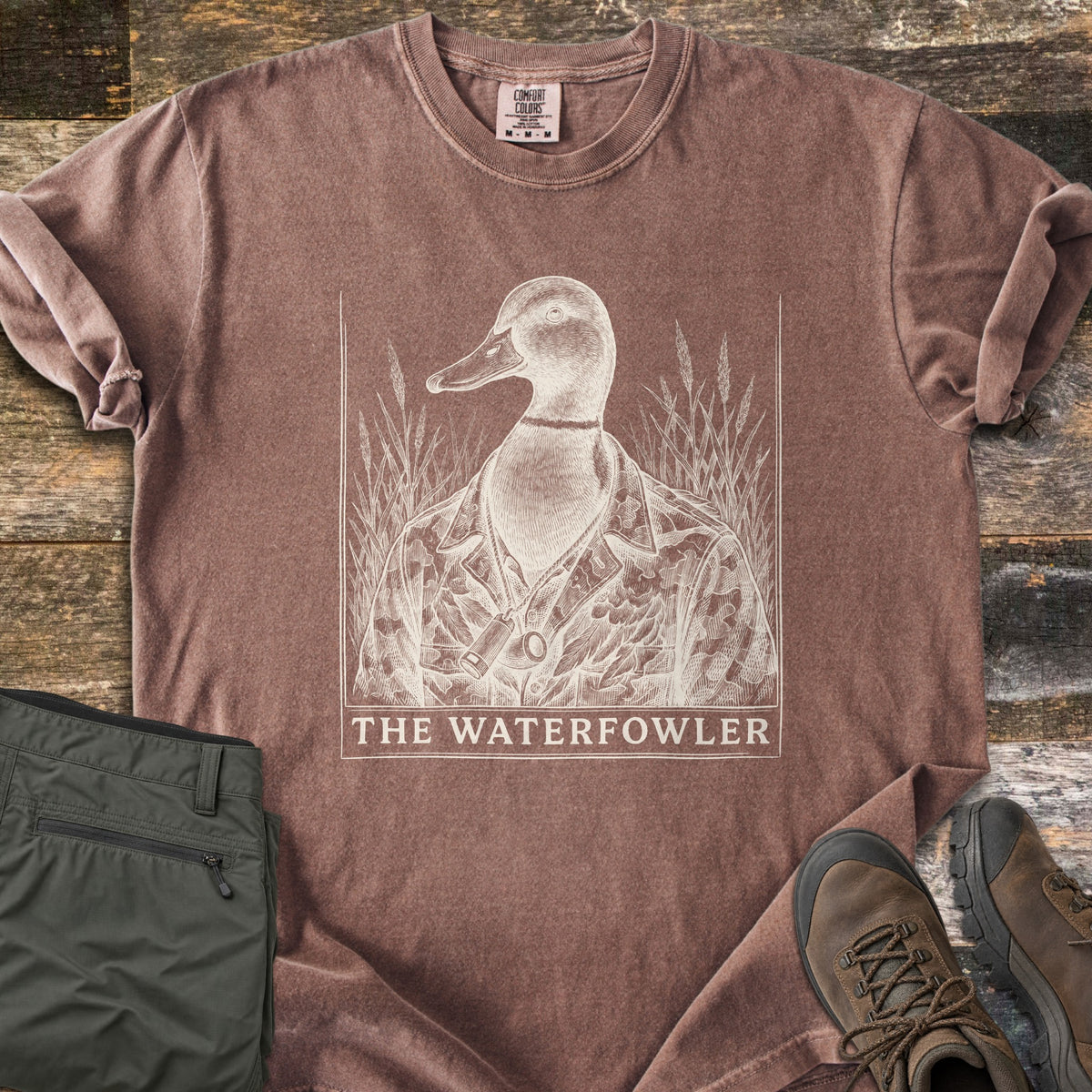 The Waterfowler Comfort Colors T-shirt in Espresso, laid flat. A cream engraving-style mallard in a camo jacket with a duck call on a cord stands in marsh grass above the words The Waterfowler. Folded olive pants and brown hiking boots sit beside it on weathered wood.