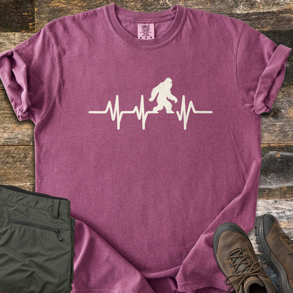 Bigfoot Heartbeat Comfort Colors T-shirt in Berry, laid flat. A cream heartbeat line runs across the chest with a walking Bigfoot silhouette in the middle; there are no words. The shirt lies on weathered wood beside folded olive hiking pants and brown hiking boots.