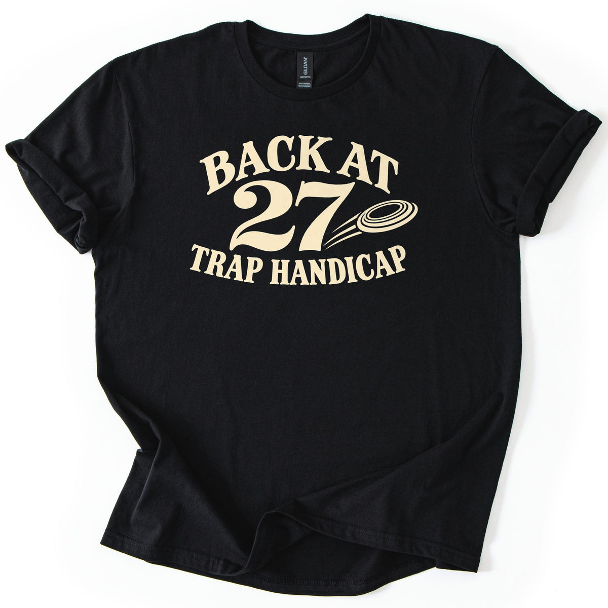 Back at 27 Trap Handicap Comfort Colors T-shirt in Black, laid flat. Cream lettering reads Back at 27 Trap Handicap, with a flying clay target and motion lines next to the large 27. The shirt has rolled sleeves and lies on a white background with its hem slightly folded.
