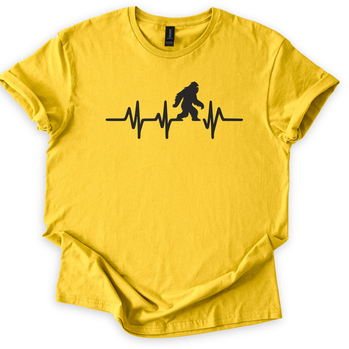 Yeti Heartbeat Comfort Colors T-shirt in Daisy, laid flat. A black heartbeat line runs across the chest, with a walking Bigfoot silhouette standing within it; there are no words. The sleeves are cuffed and the shirt lies on a white background.