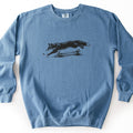 Leaping Fox Comfort Colors crewneck sweatshirt in Blue Jean, laid flat. A black ink-style illustration of a fox leaping over a low wooden rail fence and grass, with a small leaf ahead of its paws, printed across the chest. No words. The sweatshirt lies on a white surface.