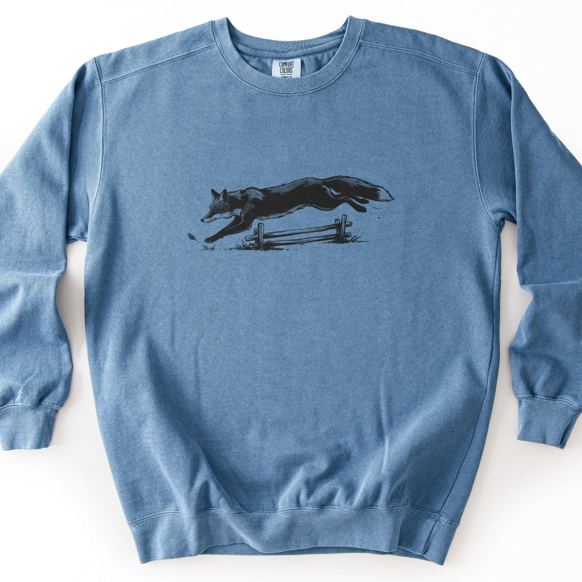 Leaping Fox Comfort Colors crewneck sweatshirt in Blue Jean, laid flat. A black ink-style illustration of a fox leaping over a low wooden rail fence and grass, with a small leaf ahead of its paws, printed across the chest. No words. The sweatshirt lies on a white surface.