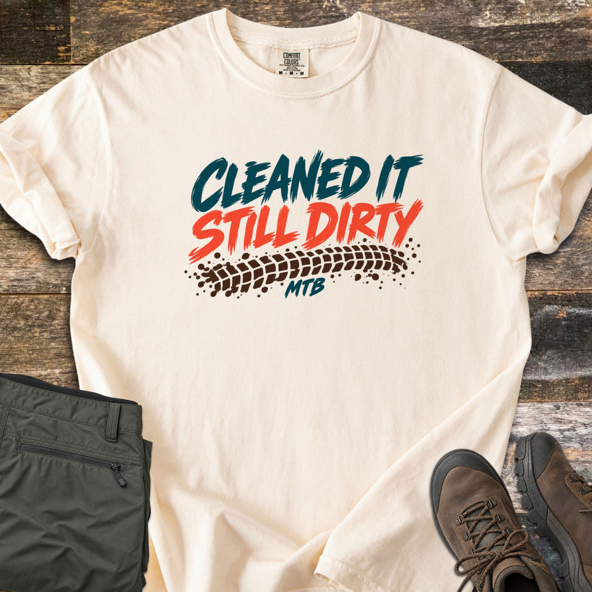 Cleaned It Still Dirty Comfort Colors T-shirt in Ivory, laid flat. Brushstroke lettering reads Cleaned It in teal and Still Dirty in orange above a muddy brown tire track and the word MTB. Olive hiking pants and brown hiking boots sit beside it on weathered wood planks.