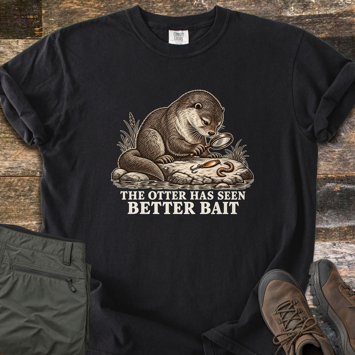 The Otter Has Seen Better Bait Comfort Colors T-shirt in Black, laid flat. A detailed brown illustration of an otter on a rock studying a fishing lure and a worm through a magnifying glass, above the words The Otter Has Seen Better Bait in cream. Olive hiking pants and brown boots lie on wood planks.