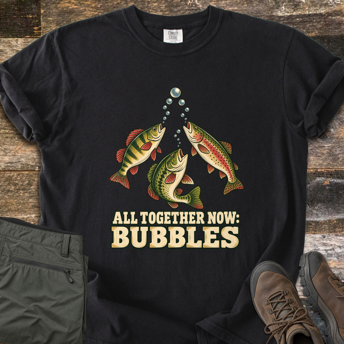All Together Now: Bubbles Comfort Colors T-shirt in Black, laid flat. Three fish, a yellow perch, a largemouth bass and a rainbow trout, blow rising bubbles above the words All Together Now: Bubbles. Olive pants and brown hiking boots lie beside it on weathered wood boards.