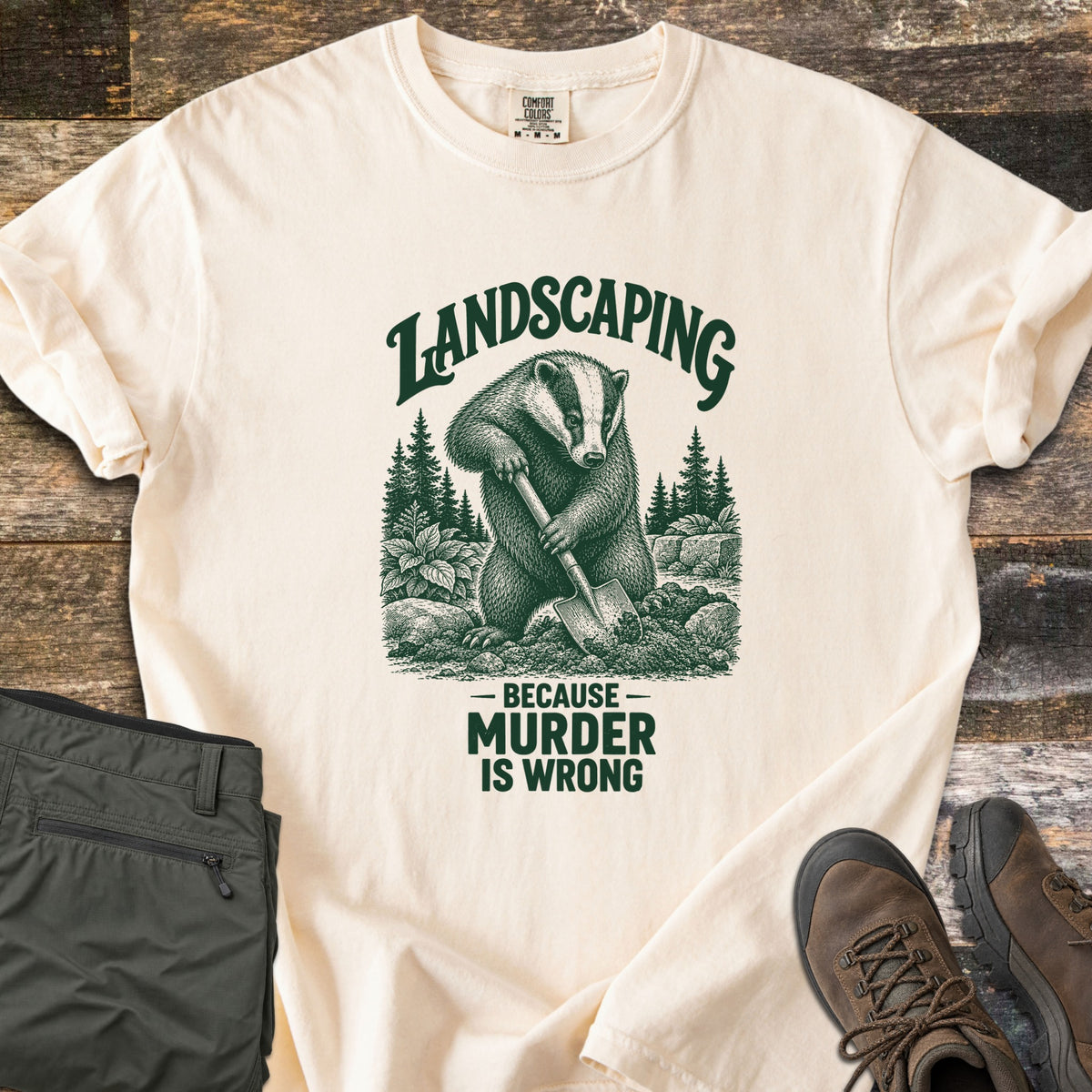 Landscaping Because Murder Is Wrong Comfort Colors T-shirt in Ivory, laid flat. A dark green engraving-style badger digs with a shovel among pine trees, plants and rocks under the arched word Landscaping, above Because Murder Is Wrong. Set on weathered wood beside olive hiking pants and brown boots.