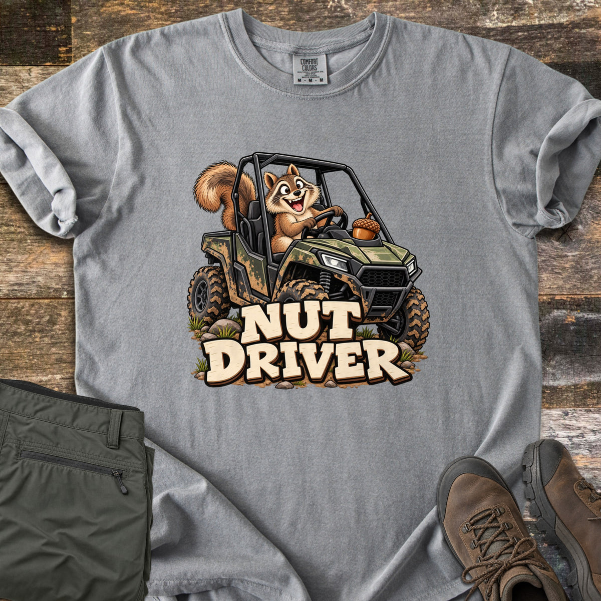 Nut Driver Comfort Colors T-shirt in Grey, laid flat. A cartoon squirrel grins while driving a muddy camo side-by-side with an acorn on the hood, above the words Nut Driver. Olive cargo pants and brown hiking boots sit beside it on weathered wood.