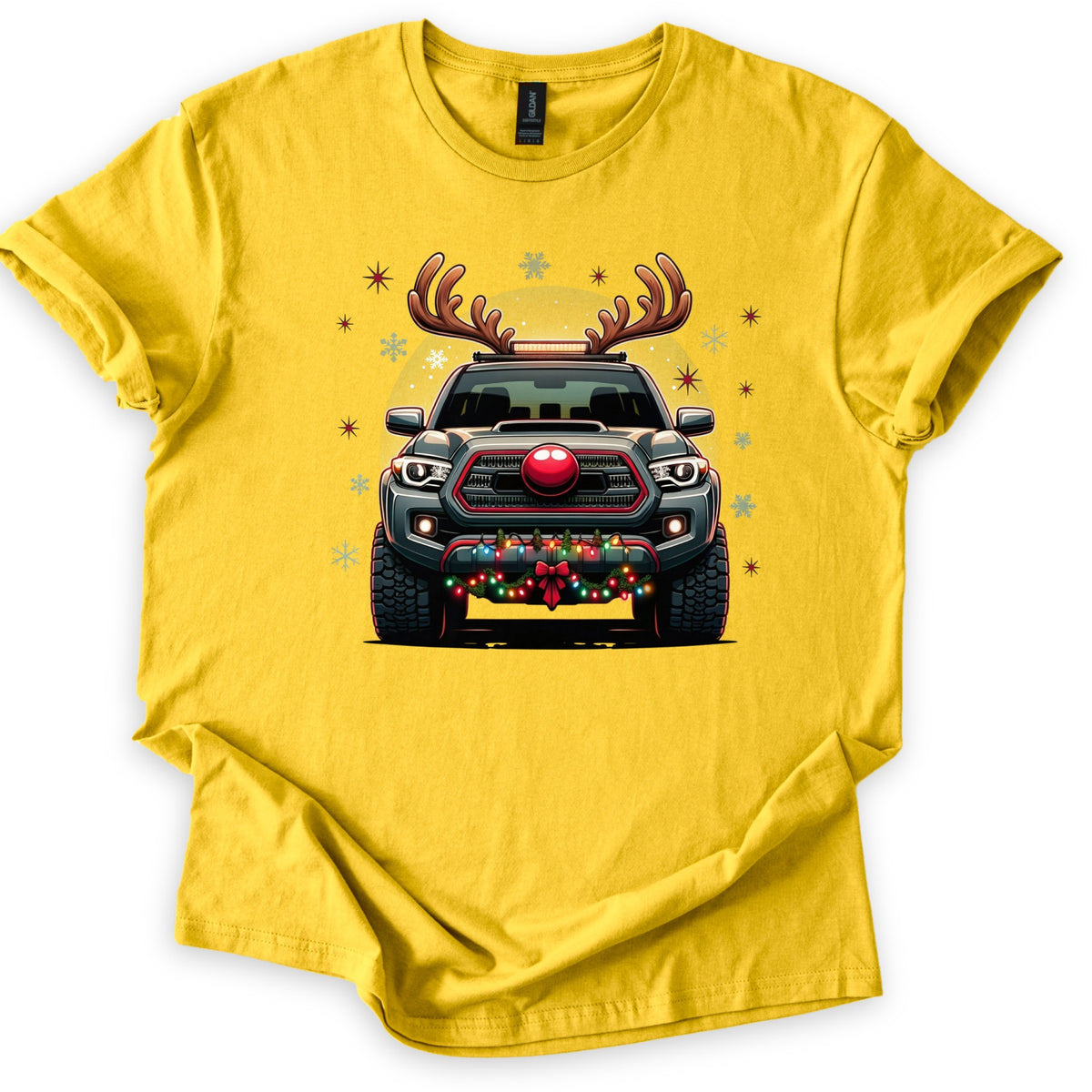 Santa Truck Comfort Colors T-shirt in Daisy, laid flat. A dark gray pickup truck seen head-on wears reindeer antlers and a red nose, with colored string lights and a red bow across its bumper, surrounded by snowflakes and red stars. No words. White background.
