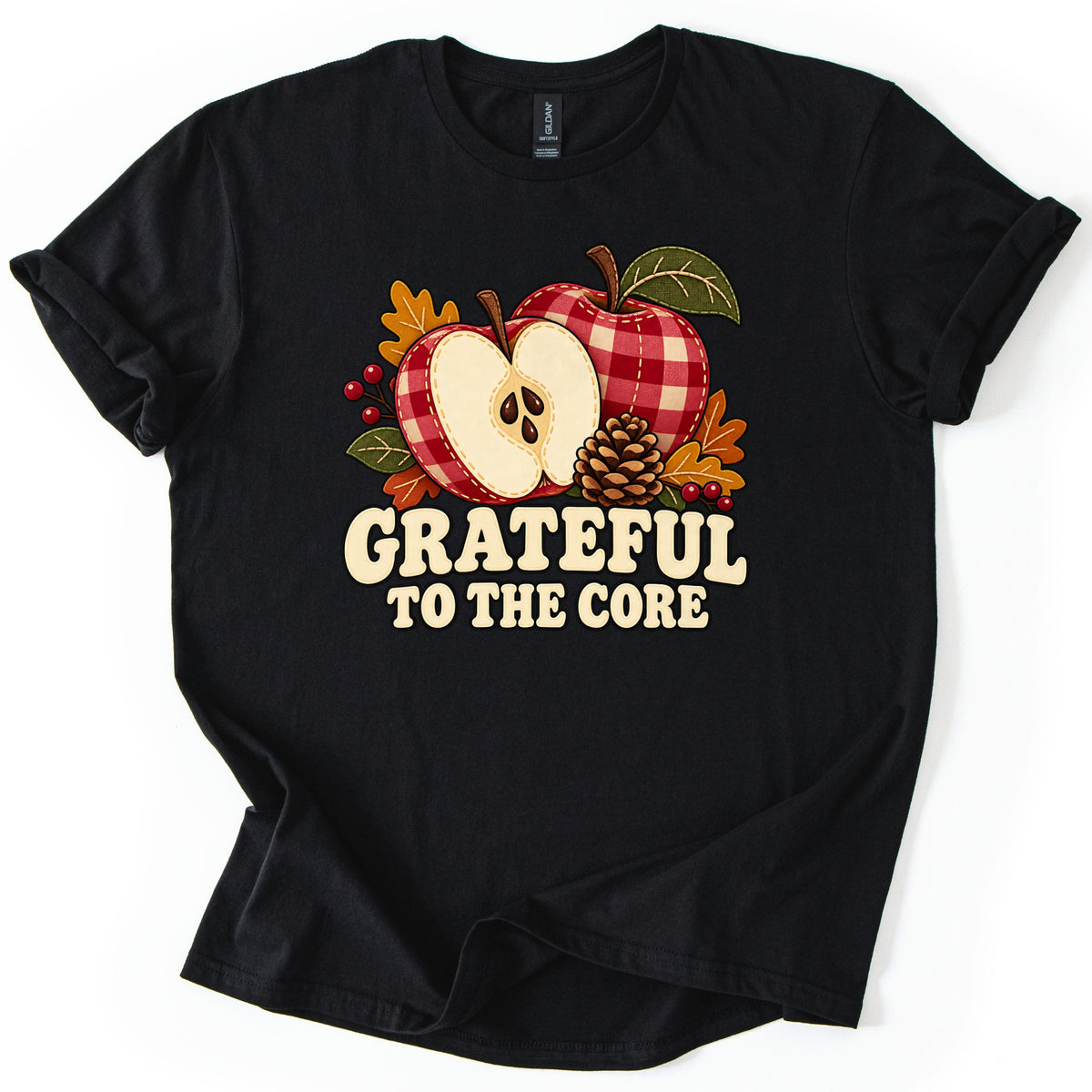 Grateful to the Core Comfort Colors T-shirt in Black, laid flat. A whole red gingham apple and a halved one showing seeds, with a pinecone, fall leaves and berries, sit above cream letters reading Grateful to the Core. The shirt lies on a white surface with cuffed sleeves.