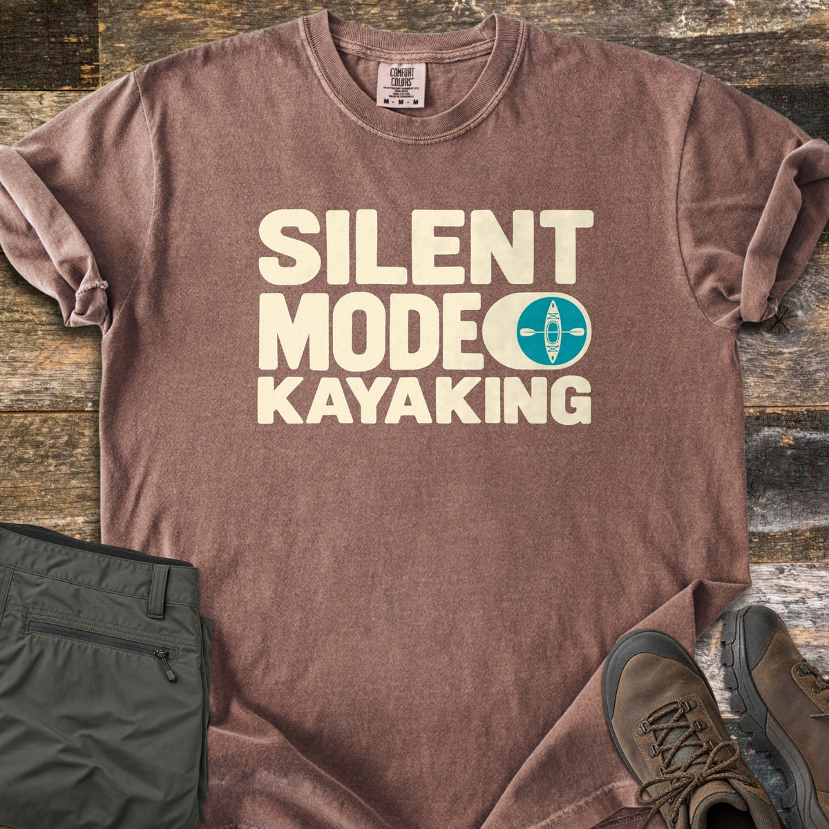 Silent Mode Comfort Colors T-shirt in Espresso, laid flat. Cream block letters read SILENT MODE KAYAKING, with a toggle switch icon showing a teal circle with a kayak and paddle. Sleeves are rolled on a wood background beside folded olive pants and brown hiking boots.