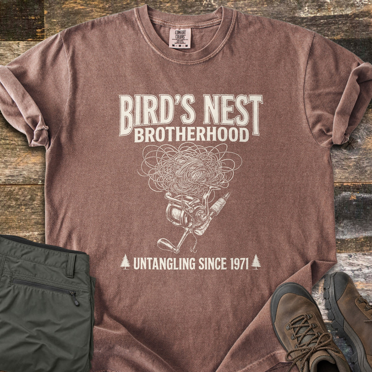 Birds Nest Brotherhood Comfort Colors T-shirt in Espresso, laid flat. Cream sketch of a tangled line nest over a spinning reel, rod and hook, with Bird's Nest Brotherhood above and Untangling Since 1971 between pine trees below. Olive pants and hiking boots lie on old wood boards.