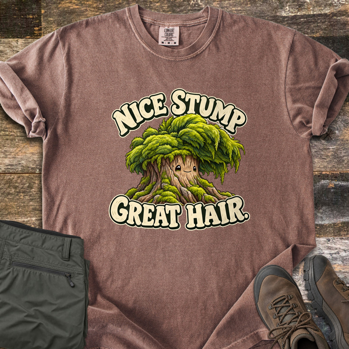 Nice Stump Great Hair Comfort Colors T-shirt in Espresso, laid flat. A smiling cartoon tree stump with a shaggy mop of green moss hair sits between the words Nice Stump Great Hair. Sleeves rolled, on weathered wood with folded olive hiking pants and brown hiking boots.