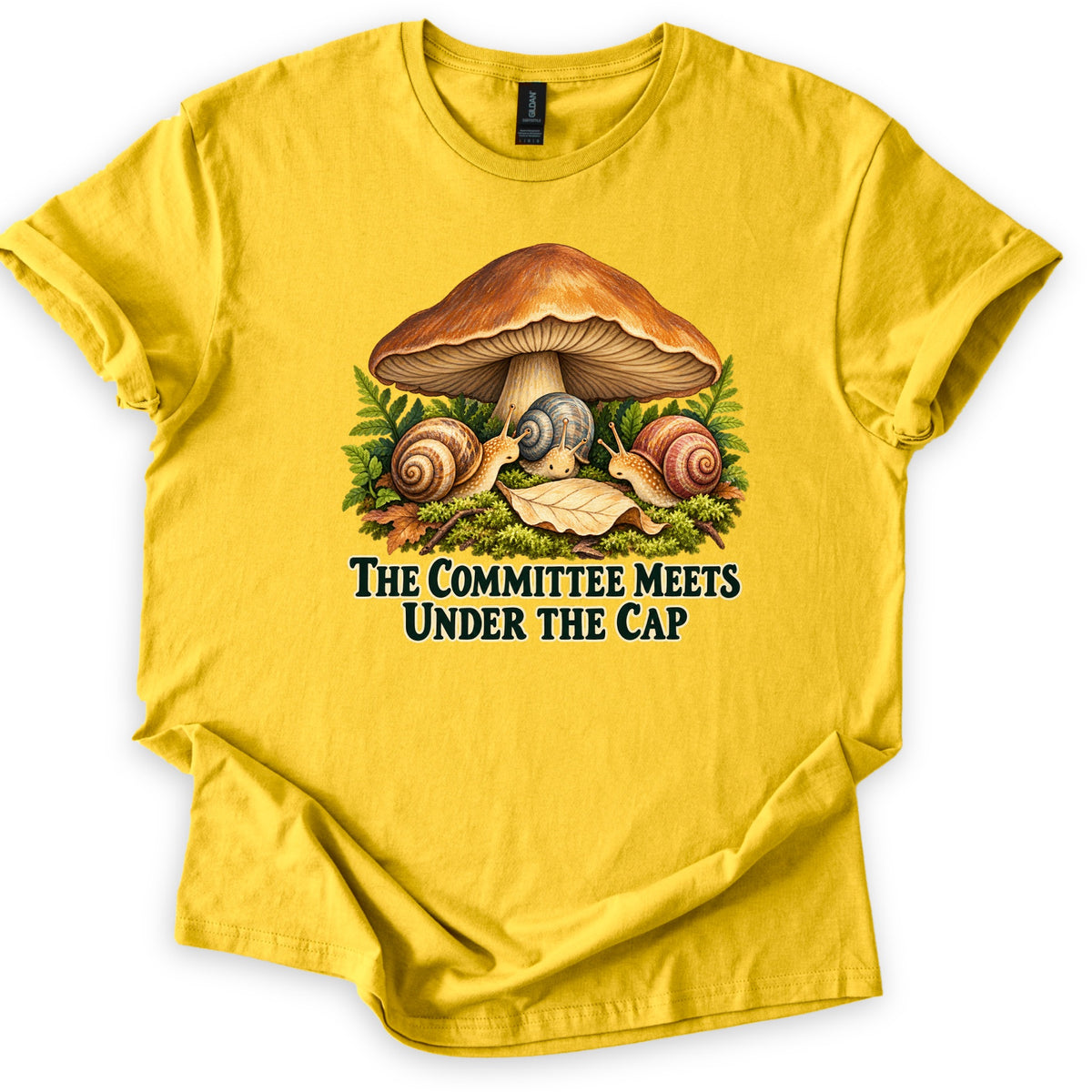The Committee Meets Under the Cap Comfort Colors T-shirt in Daisy, laid flat. Three snails gather on moss beneath a large brown mushroom, framed by ferns and a fallen leaf, above dark green words The Committee Meets Under the Cap. The shirt lies on a white background with rolled sleeves.