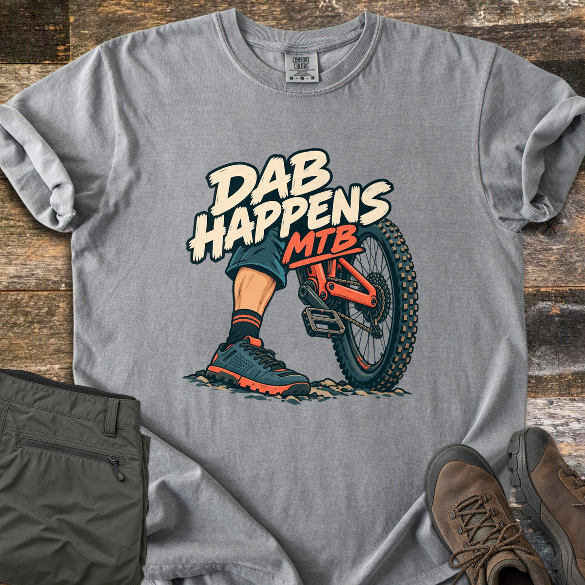 Dab Happens Comfort Colors T-shirt in Grey, laid flat. A cartoon rider's foot in a sneaker is planted on rocky dirt beside an orange mountain bike's rear wheel, under the words Dab Happens MTB. Olive pants and brown hiking boots sit on a weathered wood surface.