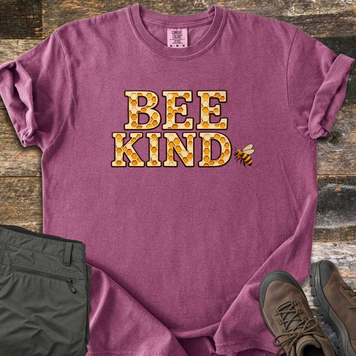 Bee Kind Comfort Colors T-shirt in Berry, laid flat. The words BEE KIND in large letters filled with a golden honeycomb pattern, with a small bee beside them. Sleeves are rolled, on a weathered wood floor beside folded olive cargo pants and brown hiking boots.