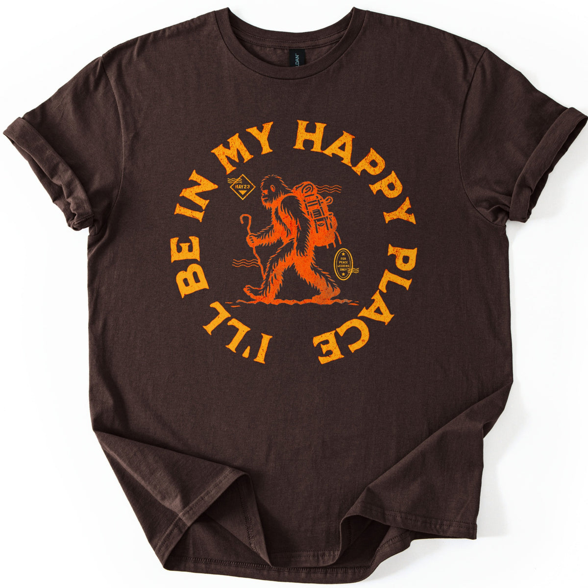 Happy Place Hiking Squatch Comfort Colors T-shirt in Dark Chocolate, laid flat. An orange Bigfoot with a backpack and walking stick strides inside a circle of the words I'll Be In My Happy Place, next to a small oval reading For Peace Seekers Only. The shirt lies on white with cuffed sleeves.