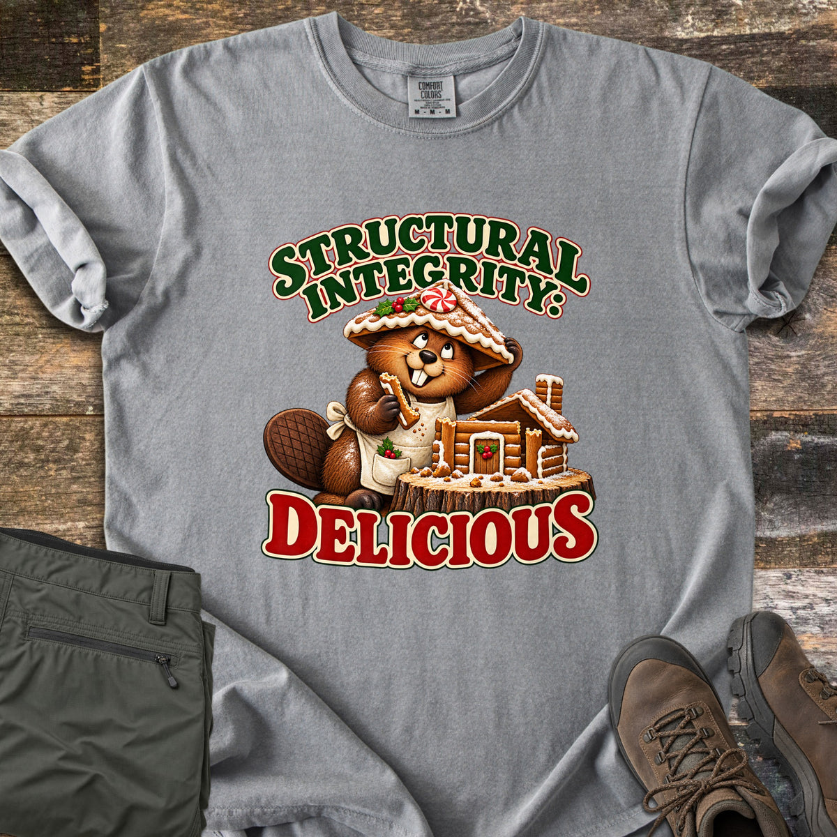 Structural Integrity: Delicious Comfort Colors T-shirt in Grey, laid flat. A cartoon beaver wearing a gingerbread roof as a hat nibbles a cookie beside a gingerbread log cabin on a tree stump, between the words Structural Integrity: Delicious. Olive pants and brown boots lie on a wood floor.