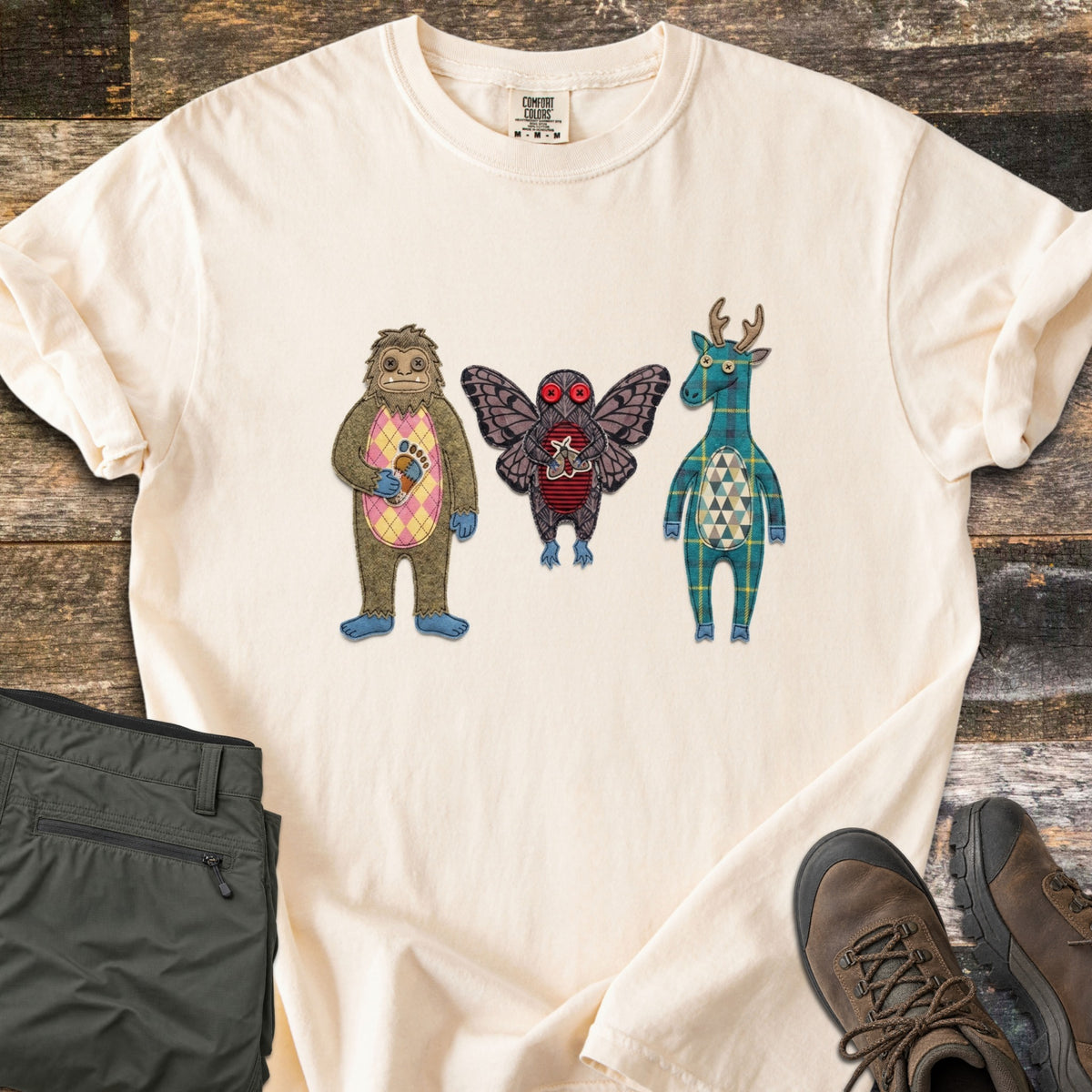 Patchwork Cryptid Trio Comfort Colors T-shirt in Ivory, laid flat. Three patchwork fabric cryptids: Bigfoot with a pink argyle belly holding a footprint patch, a red-eyed Mothman, and a plaid jackalope. Laid on weathered wood beside olive pants and brown hiking boots.