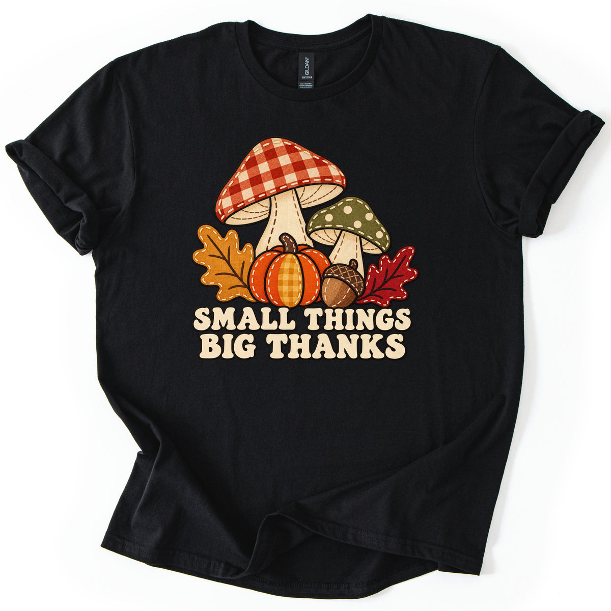 Small Things Big Thanks Comfort Colors T-shirt in Black, laid flat. Patchwork-style red gingham and green polka-dot mushrooms stand behind a plaid pumpkin, an acorn and oak leaves above the words Small Things Big Thanks. The sleeves are rolled on a white surface.