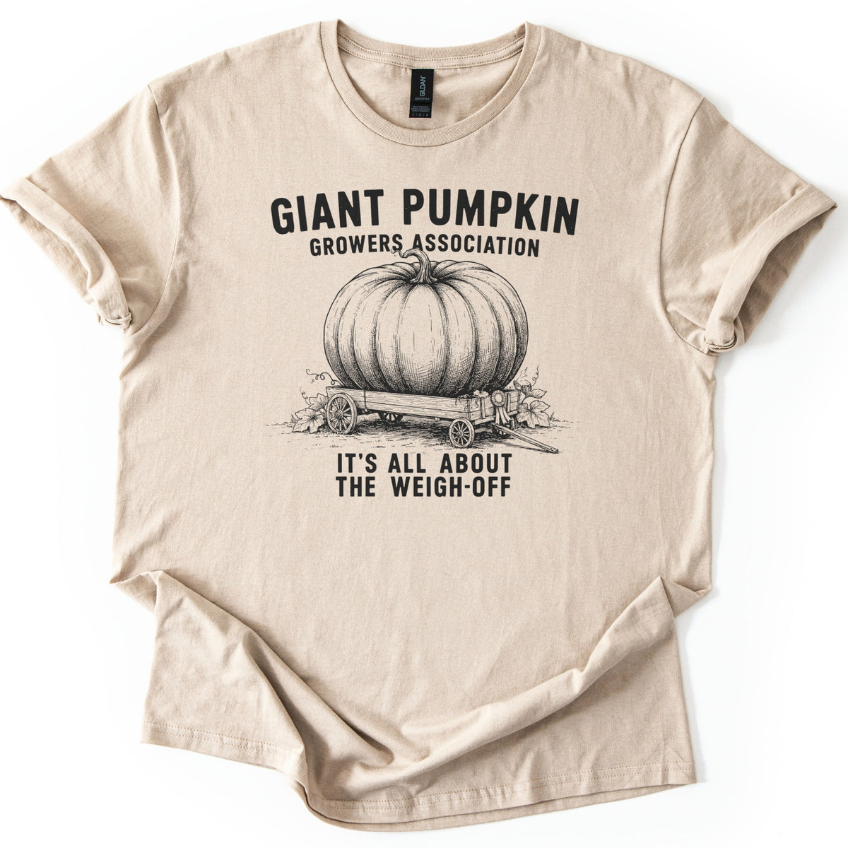 Giant Pumpkin Growers Association Comfort Colors T-shirt in Sand, laid flat. A black engraving-style drawing of a giant pumpkin on a wooden wagon with a prize ribbon sits between the words Giant Pumpkin Growers Association and It's All About the Weigh-Off. Sleeves cuffed, on white.