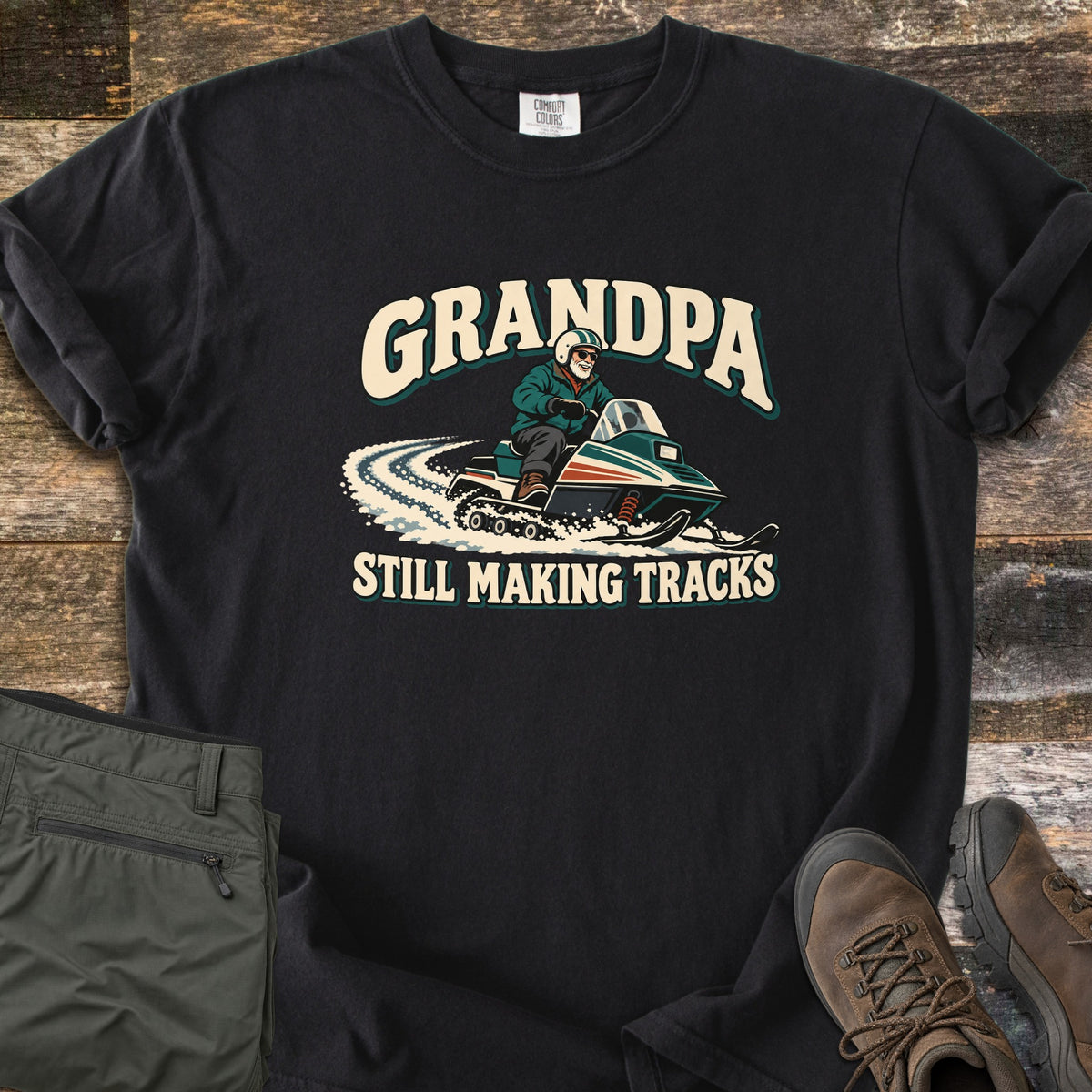 Grandpa Still Making Tracks Comfort Colors T-shirt in Black, laid flat. A cartoon grandpa in a helmet and sunglasses rides a vintage snowmobile through snow, with GRANDPA above and STILL MAKING TRACKS below. Olive pants and brown hiking boots sit beside it on weathered wood.