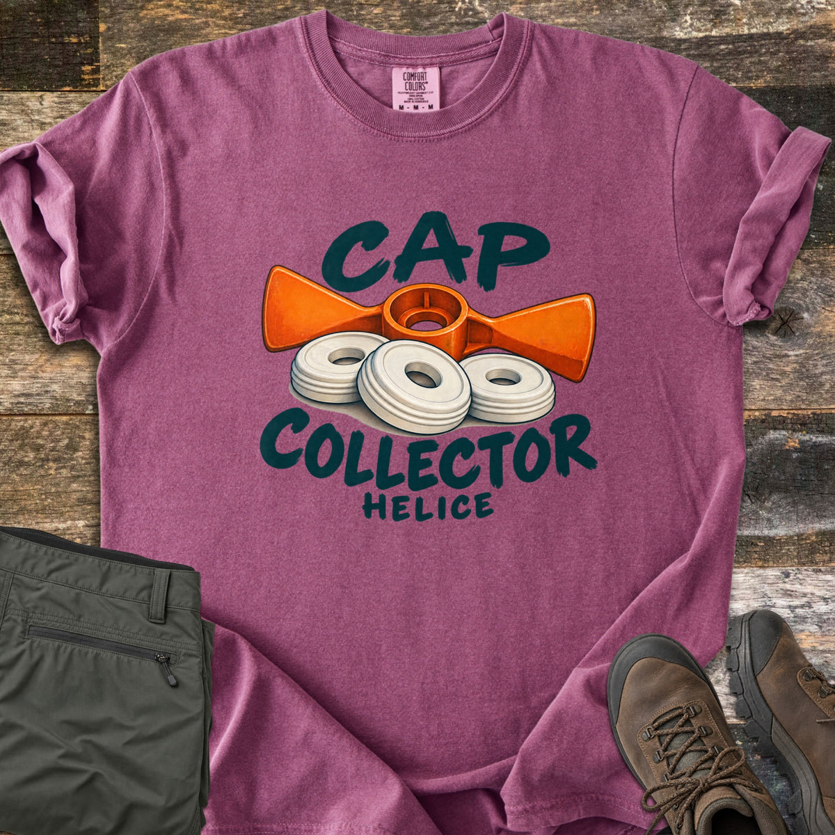 Cap Collector Comfort Colors T-shirt in Berry, laid flat. An orange helice key and three white caps sit between the words Cap Collector, with Helice below. Sleeves are rolled; olive pants and brown hiking boots lie beside it on dark wood planks.