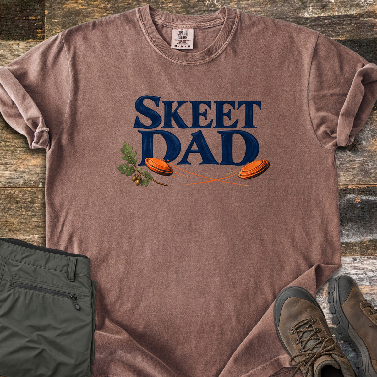 Skeet Dad Comfort Colors T-shirt in Espresso, laid flat. Navy text reads Skeet Dad above two orange clay targets with motion trails and an oak sprig with acorns. Sleeves are rolled on a wood floor beside folded olive cargo pants and brown hiking boots.