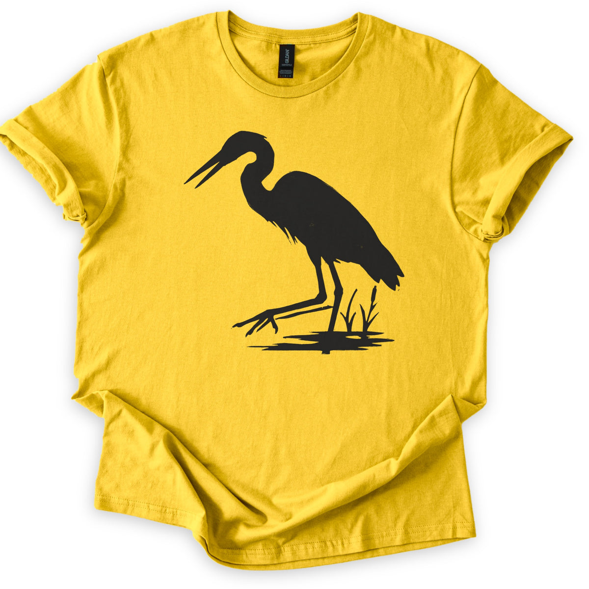 Great Blue Heron Comfort Colors T-shirt in Daisy, laid flat. A black silhouette of a heron wading in shallow water beside cattails, beak open and one foot reaching forward. The shirt lies on a white surface with cuffed sleeves.