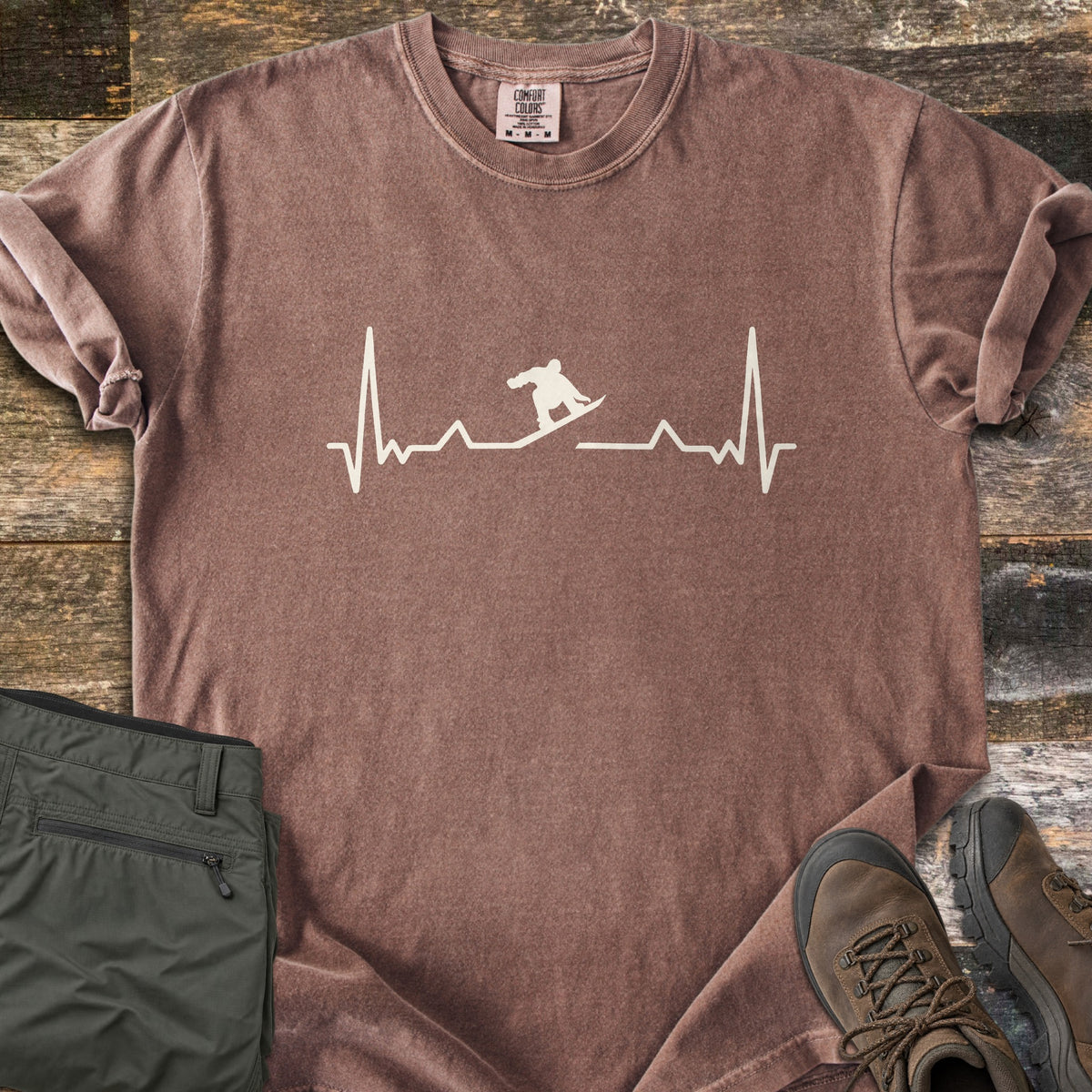 Snowboarder Heartbeat Comfort Colors T-shirt in Espresso, laid flat. A cream heartbeat line runs across the chest, with a snowboarder silhouette riding the line in the middle. Sleeves are rolled, on a rustic wood floor beside folded olive pants and brown hiking boots.