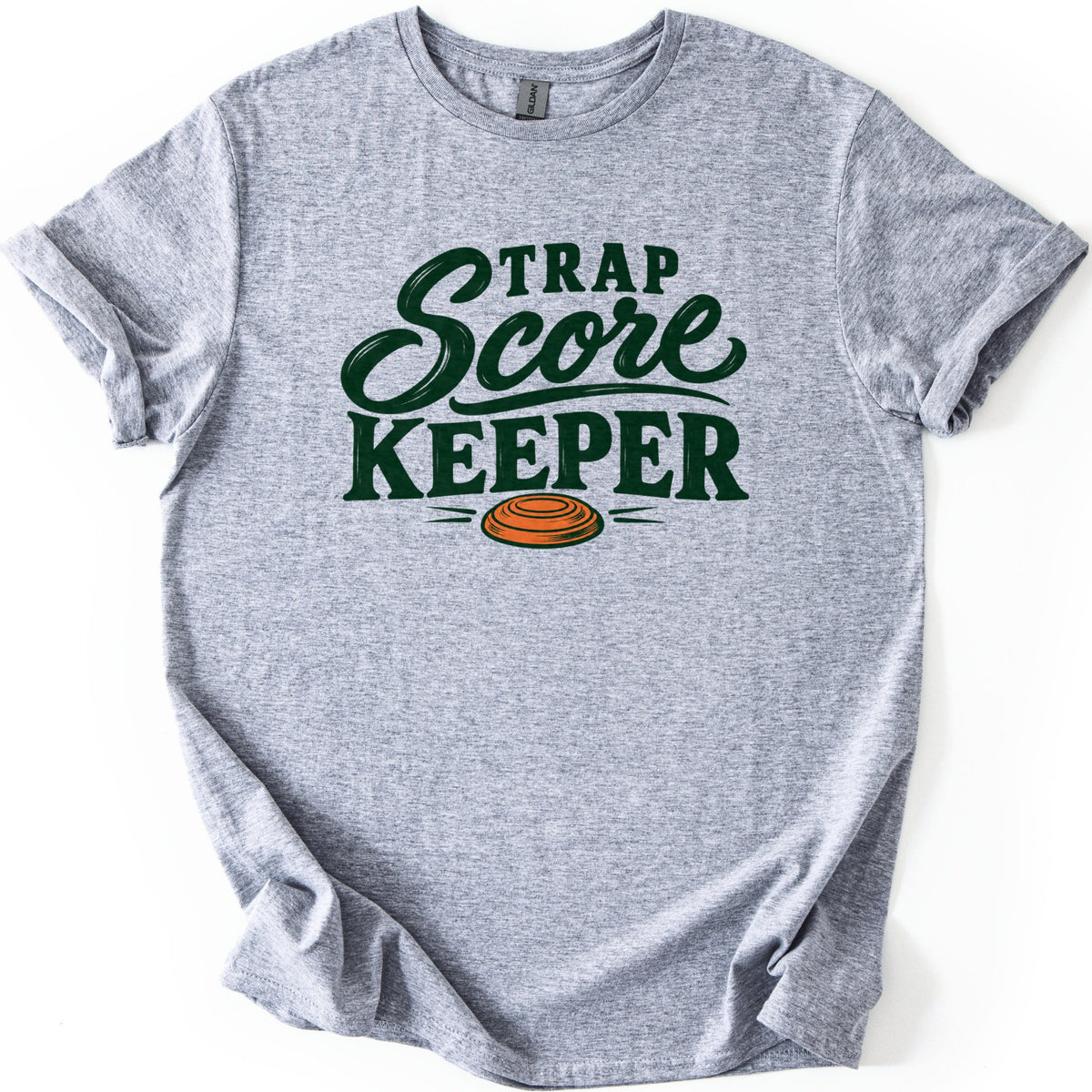 Trap Score Keeper Comfort Colors T-shirt in Sport Grey, laid flat. Dark green lettering reads Trap Score Keeper, with Score in flowing script above an orange clay target with motion lines. The sleeves are cuffed and it lies on a plain white background.