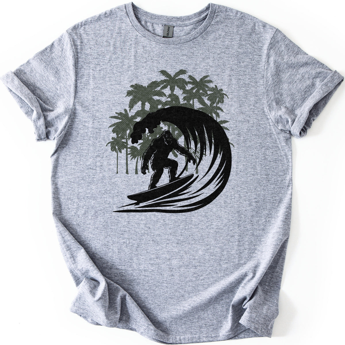 Surf Squatch Comfort Colors T-shirt in Sport Grey, laid flat. A black silhouette of Bigfoot riding a surfboard through a curling wave, with olive green halftone palm trees behind him. No words are printed. The shirt has rolled sleeves on a white background.