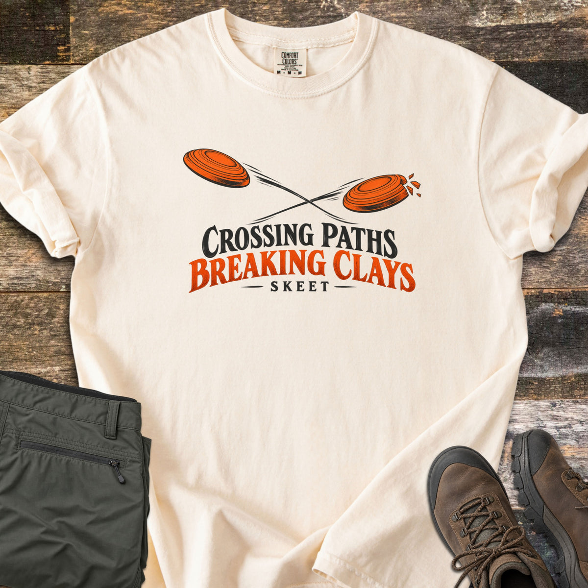 Crossing Paths Breaking Clays Comfort Colors T-shirt in Ivory, laid flat. Two orange clay targets cross paths, one shattering, above the words Crossing Paths Breaking Clays and Skeet. Sleeves rolled, on dark barn wood with olive pants and brown hiking boots.