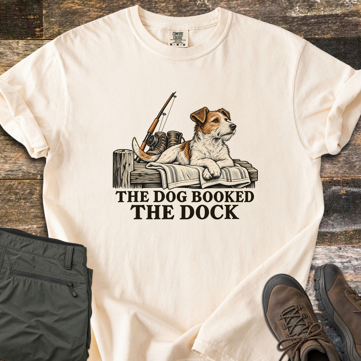 The Dog Booked the Dock Comfort Colors T-shirt in Ivory, laid flat. An illustrated Jack Russell terrier lies on a towel on a wooden dock beside a fishing rod and boots, above the words The Dog Booked The Dock in black. Olive pants and brown hiking boots sit on weathered wood.