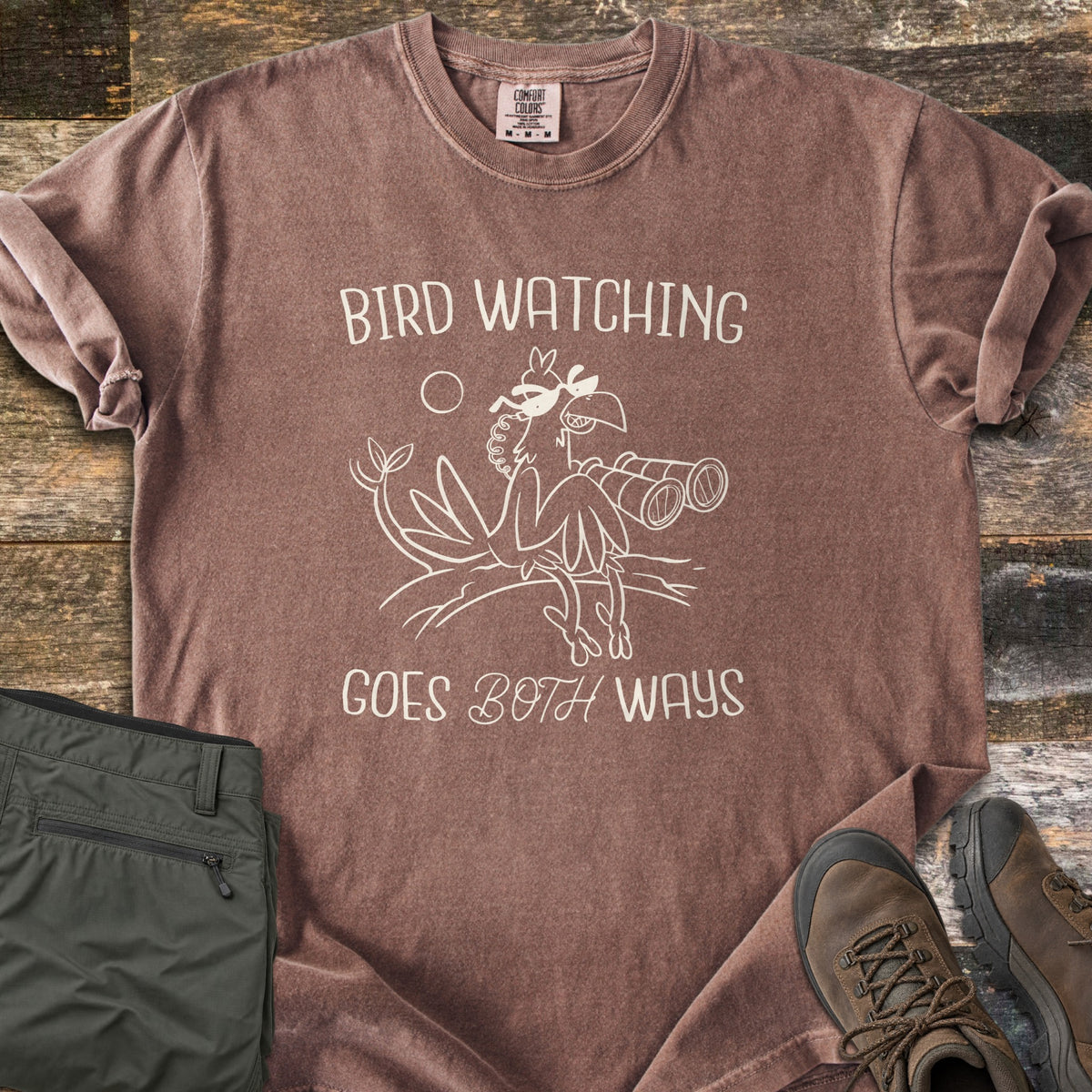 Birds Watch Back Birder Comfort Colors T-shirt in Espresso, laid flat. A cartoon bird in sunglasses perches on a branch beside binoculars between the words Bird Watching Goes Both Ways in cream. Olive hiking pants and brown hiking boots sit beside it on weathered wood.