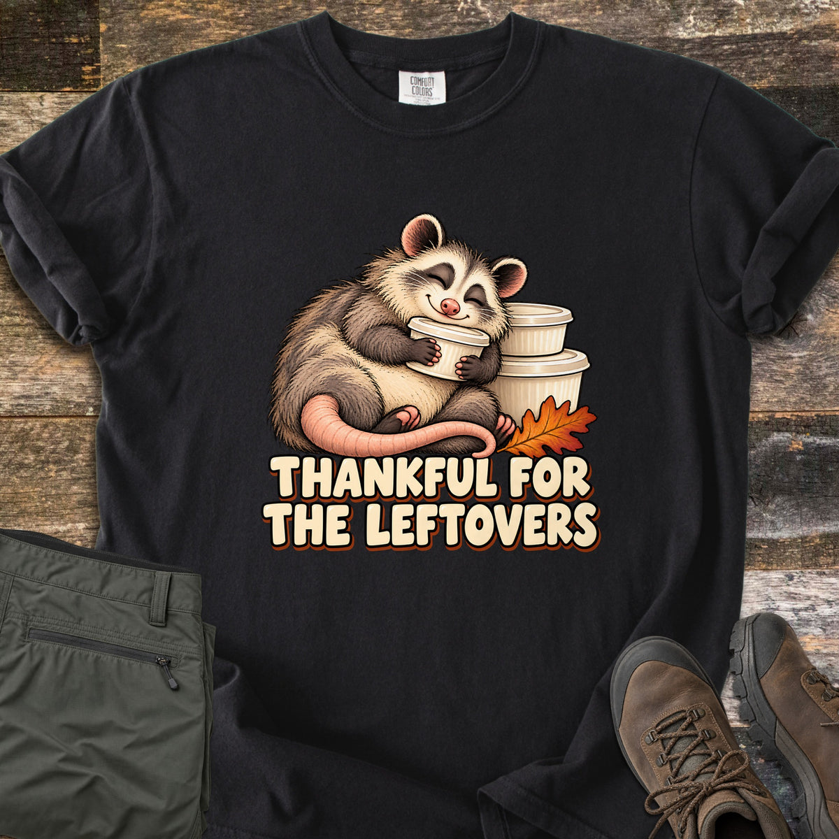 Thankful For The Leftovers Comfort Colors T-shirt in Black, laid flat. A smiling cartoon opossum hugs a white leftover container beside a stack of containers and an orange autumn leaf, above the words Thankful For The Leftovers. Olive pants and brown boots sit on a wooden floor.