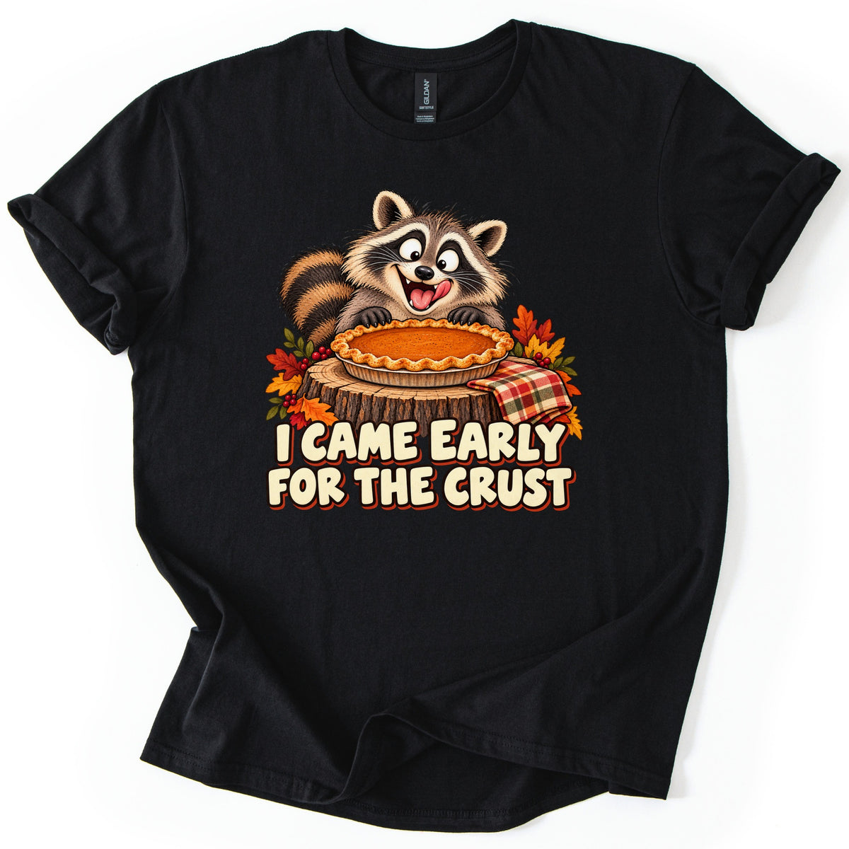 I Came Early for the Crust Comfort Colors T-shirt in Black, laid flat. A cartoon raccoon with its tongue out eyes a pumpkin pie on a tree stump, framed by fall leaves, berries and a plaid napkin, above the words I CAME EARLY FOR THE CRUST. Shown on a white background.