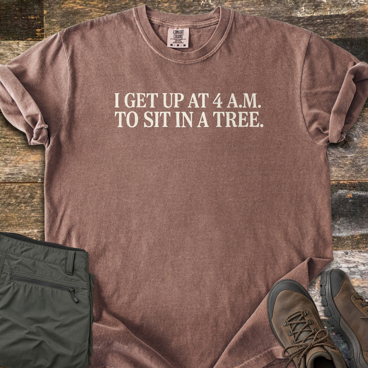 Sit In A Tree Comfort Colors T-shirt in Espresso, laid flat. Cream serif text reads I GET UP AT 4 A.M. TO SIT IN A TREE. across the chest. The shirt lies on weathered wood planks beside folded olive green cargo pants and brown leather hiking boots.