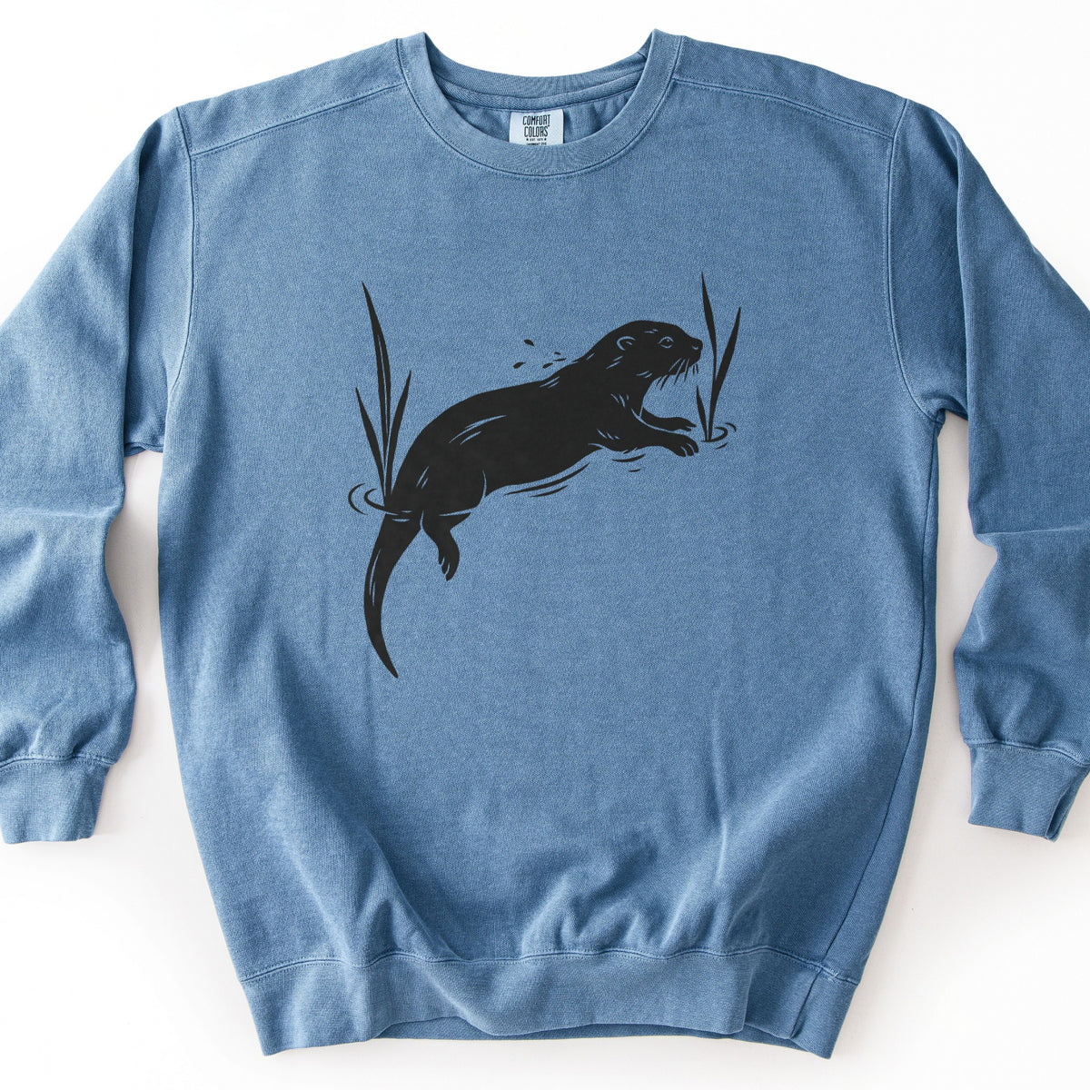 Swimming Otter Comfort Colors crewneck sweatshirt in Blue Jean, laid flat. A black block-print style otter swims with its head raised, splashing droplets, between two tall reeds with ripples around them. No words are printed. The sweatshirt lies on a white background.