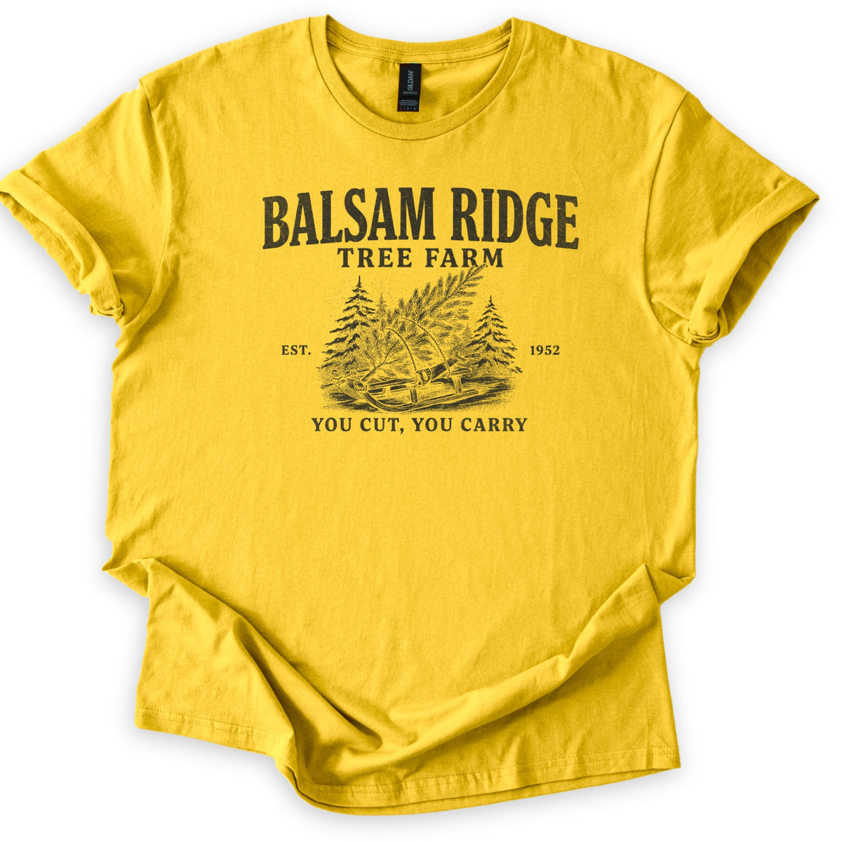 Balsam Ridge Tree Farm Comfort Colors T-shirt in Daisy, laid flat. Black print reads Balsam Ridge Tree Farm above an engraved-style fir tree tied to a wooden sled between pines, flanked by Est. 1952, with You Cut, You Carry below. Sleeves cuffed on a white surface.