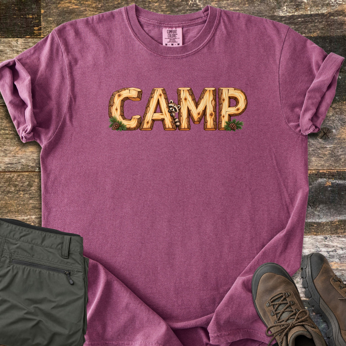 Camp Comfort Colors T-shirt in Berry, laid flat. The word CAMP is printed in letters made of cut wooden logs, with a raccoon peeking out between the A and M and pine sprigs with pinecones at each end. Olive pants and brown hiking boots sit on weathered wood planks.