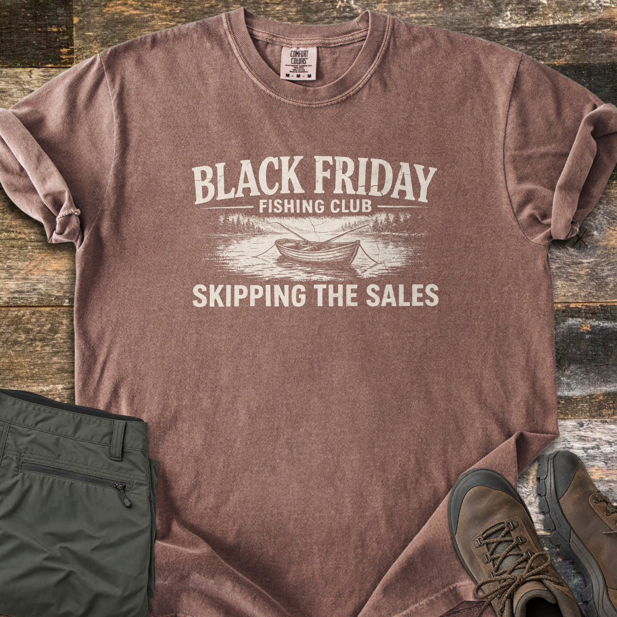 Black Friday Fishing Club Comfort Colors T-shirt in Espresso, laid flat. A distressed cream print shows the words Black Friday Fishing Club above a rowboat with oars on a lake lined with pine trees, and Skipping the Sales below. Olive pants and brown hiking boots sit beside it on wood planks.