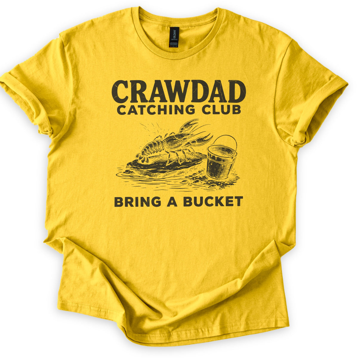 Crawdad Catching Club Comfort Colors T-shirt in Daisy, laid flat. Black print reads Crawdad Catching Club above a sketched crawdad on a flat rock at the water's edge beside a metal bucket, with Bring a Bucket below. Cuffed sleeves on a white background.