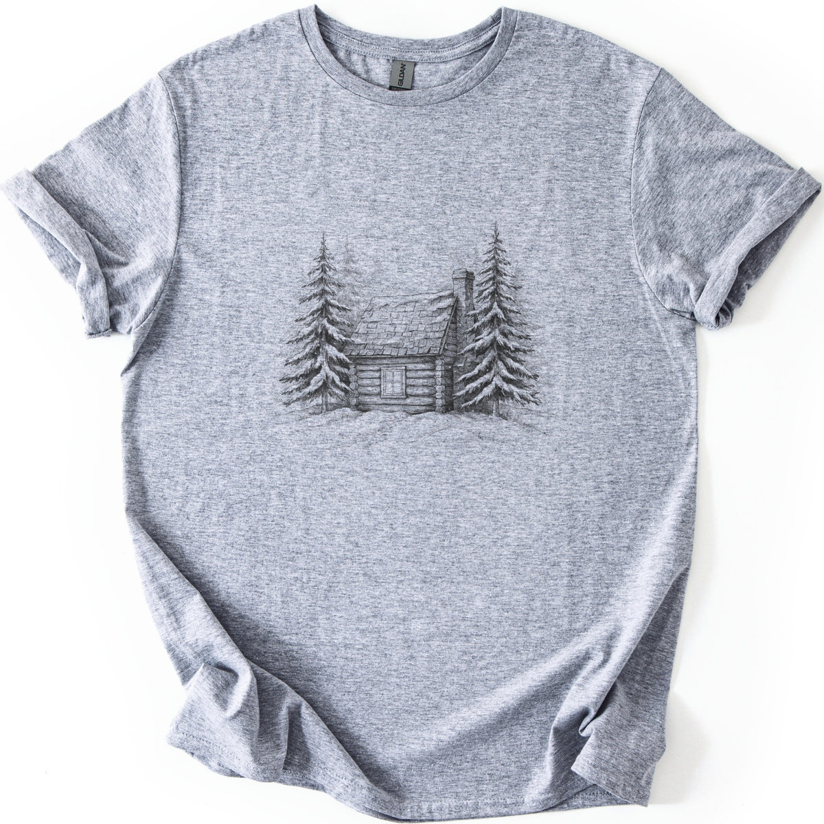 Snowy Log Cabin Comfort Colors T-shirt in Sport Grey, laid flat. A pencil-style sketch of a log cabin with a snowy roof and chimney, flanked by snow-covered pine trees on a drift. No words. Sleeves are rolled on a white background.