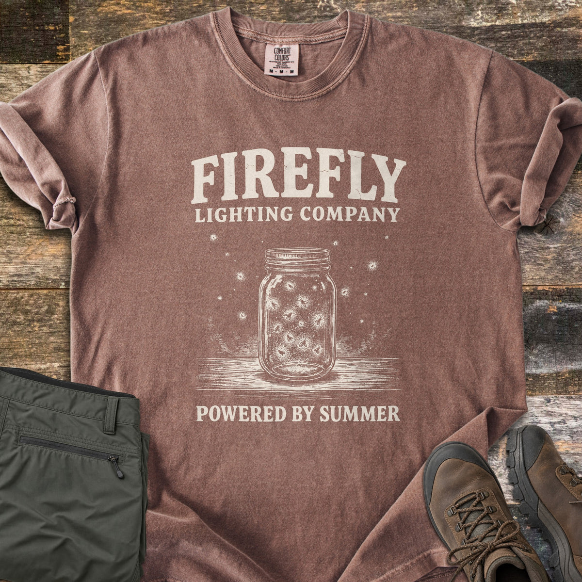 Firefly Lighting Company Comfort Colors T-shirt in Espresso, laid flat. Cream print of a mason jar full of glowing fireflies on a wooden surface, under the words Firefly Lighting Company and above Powered by Summer. Olive cargo pants and brown hiking boots lie beside it on weathered wood.