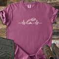 Trap Shooting Heartbeat Comfort Colors T-shirt in Berry, laid flat. A cream heartbeat line rises into a flying clay target with motion lines above three pine trees, with no words. Sleeves are rolled on weathered wood beside folded olive pants and brown hiking boots.