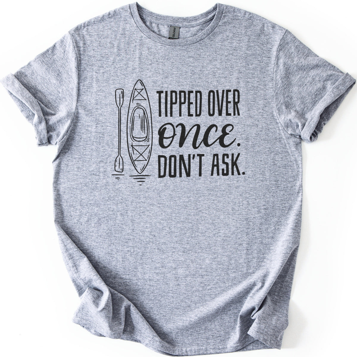 Tipped Over Once Kayak Comfort Colors T-shirt in Sport Grey, laid flat. A black line drawing of an upright kayak and paddle with water ripples sits beside the words Tipped Over Once. Don't Ask., with Once in script. The shirt has cuffed sleeves on a white background.